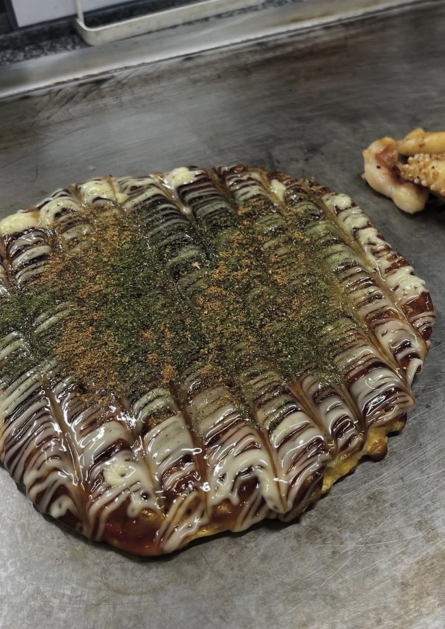 鉄板の上のお好み焼き。ソースとマヨネーズ、青のりとかつお粉がかかっている